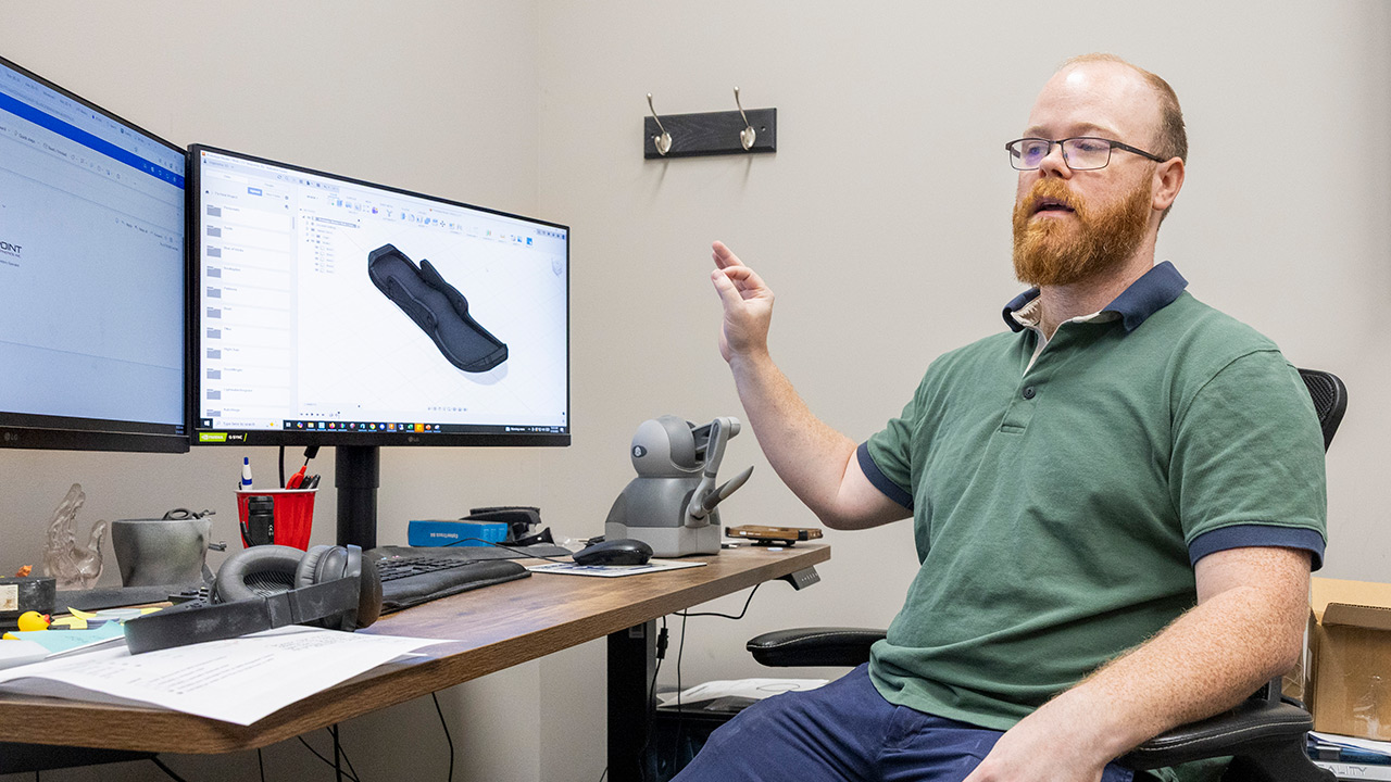 A man sits at a desk in a technical workspace, pointing at a computer monitor displaying a 3D modeling program.